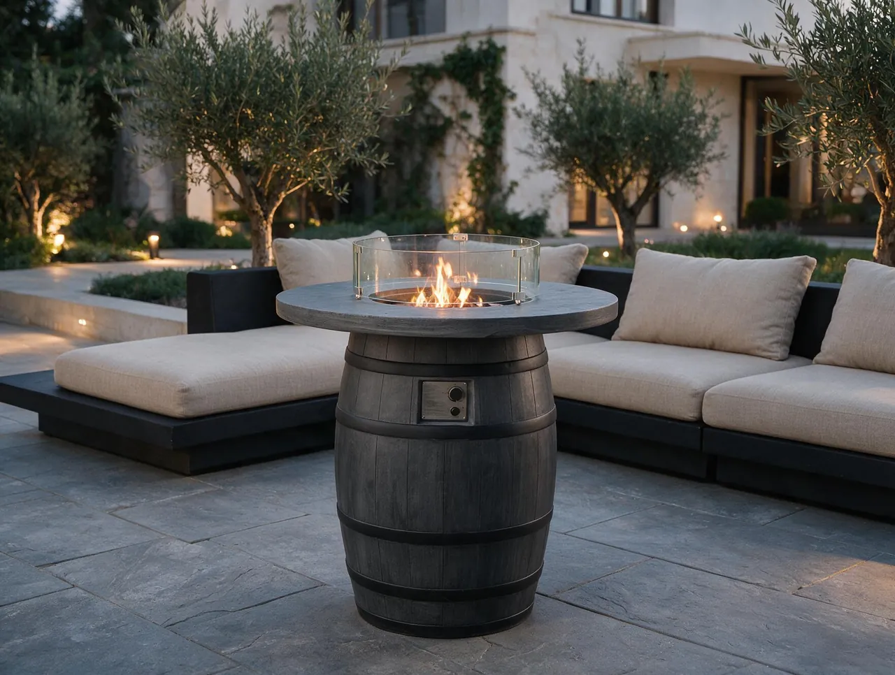 Mora Round Barrel-Shaped Gas Fire Pit Table with a glass wind guard and active flame on a stone patio near an outdoor lounge.
