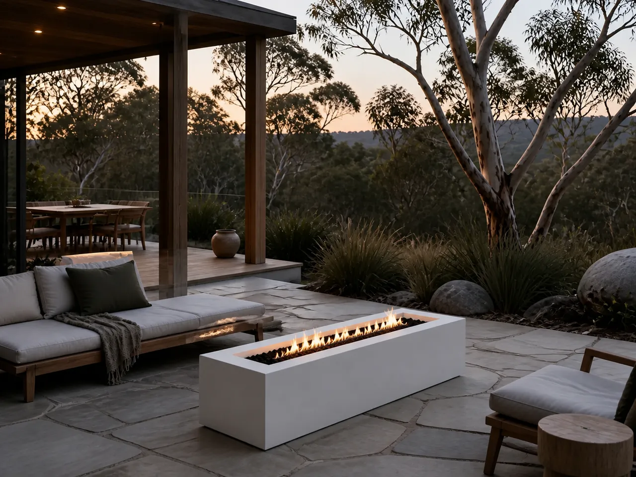 Linden Rectangular Linear Gas Fire Pit with a line of flames on a stone patio at dusk near a wooden lounge sofa.